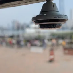 Kamera IP Avigilon 360 stopni, zamontowana na zewnątrz, z rozmytym tłem miejskim i spacerującymi ludźmi. Widoczny detal obudowy kamery.