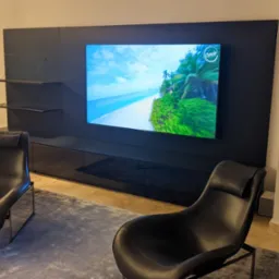 Nowoczesny salon z dużym telewizorem i czarną zabudową meblową. Na ekranie wyświetla się tropikalny widok. Dwa czarne fotele stoją na szarym dywanie.