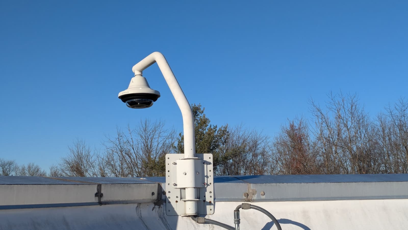 Kamera IP 360 stopni zamontowana na elewacji budynku, widoczna na tle błękitnego nieba i drzew bez liści. Biała obudowa kamery i uchwyt.