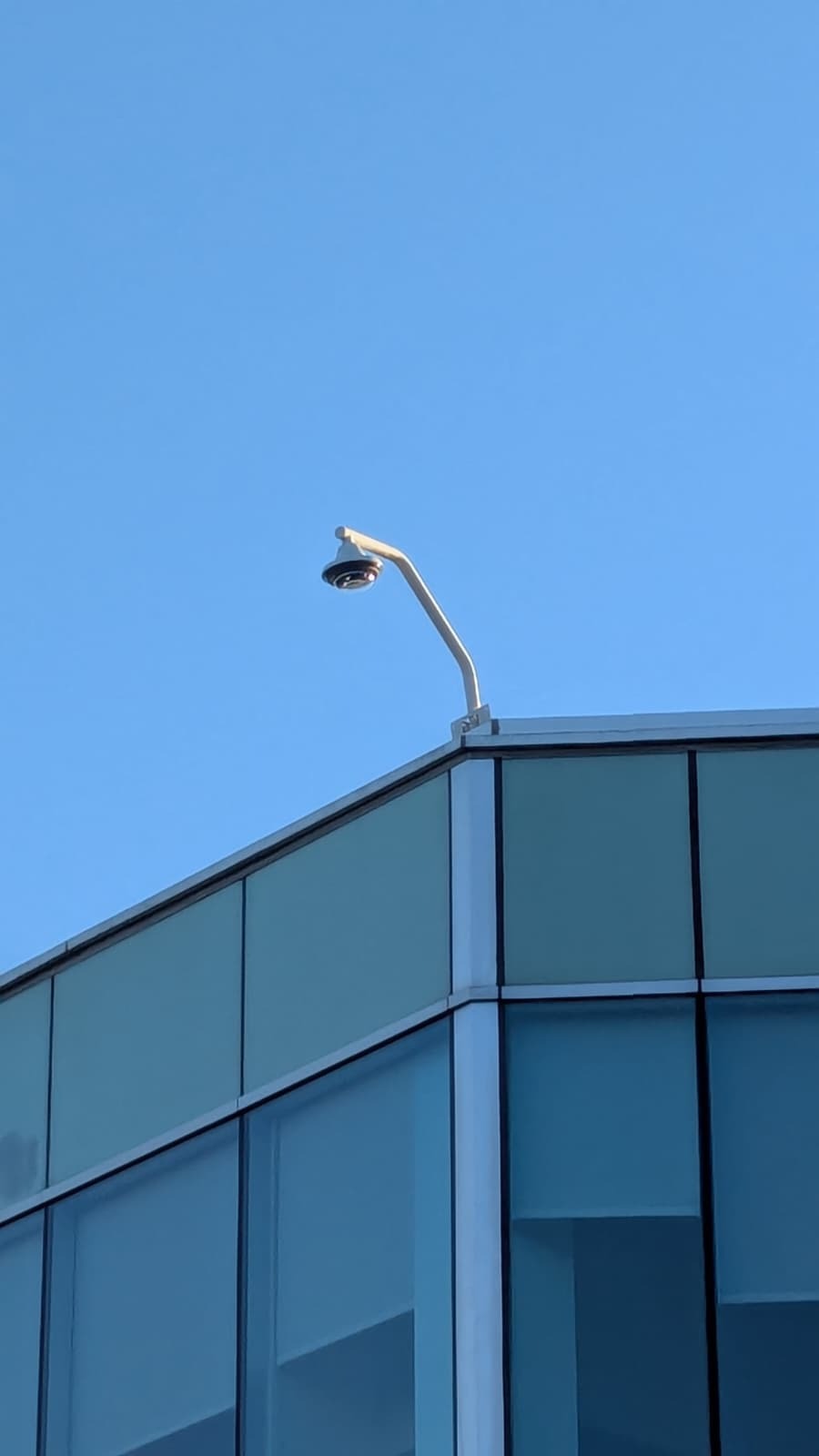 Kamera IP 360 na rogu nowoczesnego budynku biurowego z niebieskimi szybami. Ujęcie z dołu, błękitne niebo w tle. Minimalistyczny design.