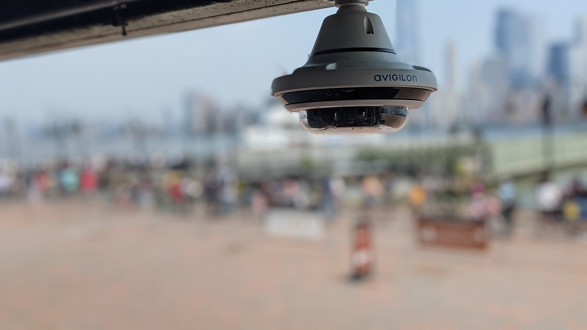 Kamera IP Avigilon 360 stopni, zamontowana na zewnątrz, z rozmytym tłem miejskim i spacerującymi ludźmi. Widoczny detal obudowy kamery.