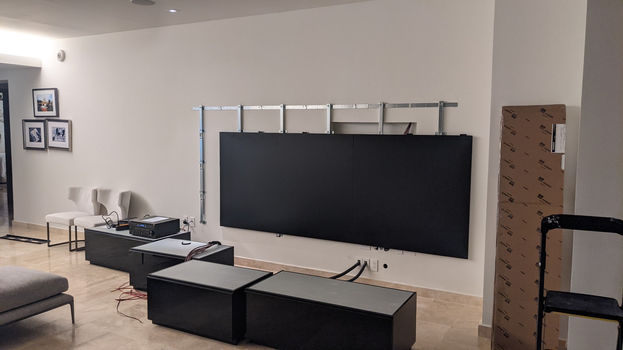 Montaż dużego ekranu TV na ścianie z widocznym systemem mocowania. Widoczne kable i sprzęt audio, etap instalacji w nowoczesnym wnętrzu.