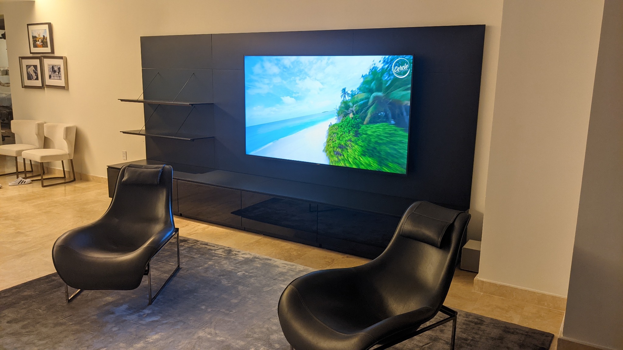 Nowoczesny salon z dużym telewizorem i czarną zabudową meblową. Na ekranie wyświetla się tropikalny widok. Dwa czarne fotele stoją na szarym dywanie.