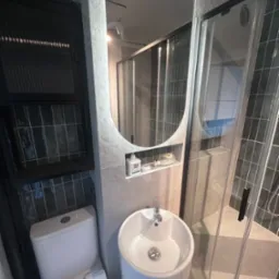 Nowoczesna łazienka z owalnym lustrem, białą umywalką na postumencie i kabiną prysznicową. Ciemne płytki na ścianach i zabudowa nad toaletą. Minimalistyczny design.