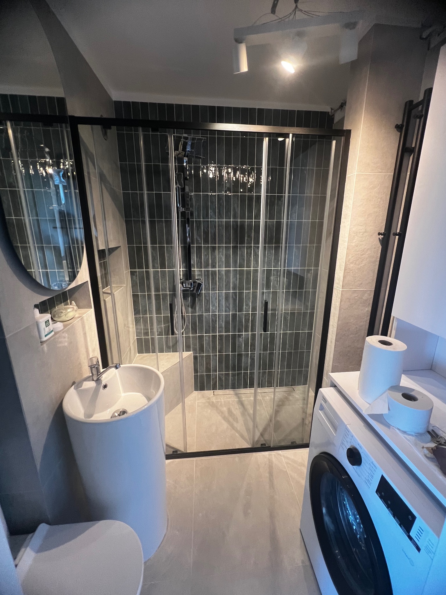 Nowoczesna łazienka z kabiną prysznicową typu walk-in, umywalką na postumencie i pralką. Szare płytki i czarne akcenty tworzą elegancki wystrój.