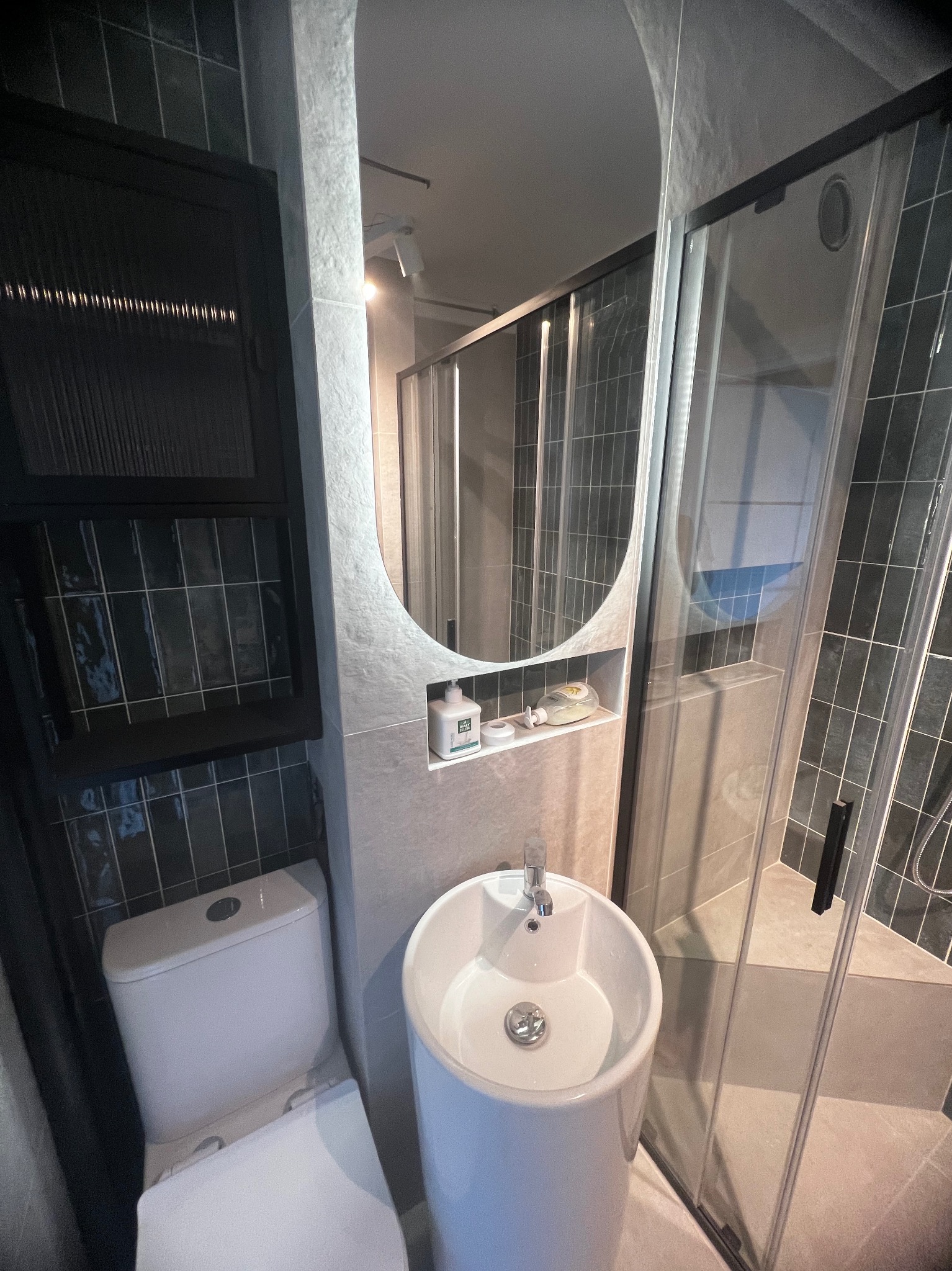 Nowoczesna łazienka z owalnym lustrem, białą umywalką na postumencie i kabiną prysznicową. Ciemne płytki na ścianach i zabudowa nad toaletą. Minimalistyczny design.