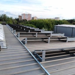 TC EnergyPro - Montaż paneli słonecznych na dachu z blachy trapezowej. Widoczne elementy konstrukcyjne i częściowo zainstalowane panele. W tle zabudowa miejska i zieleń.