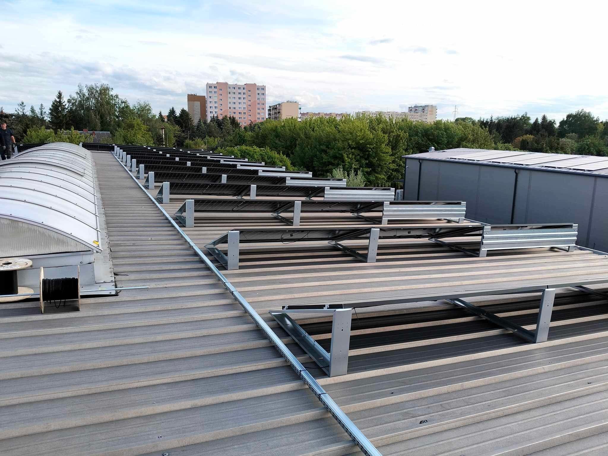 Montaż paneli słonecznych na dachu z blachy trapezowej. Widoczne elementy konstrukcyjne i częściowo zainstalowane panele. W tle zabudowa miejska i zieleń.