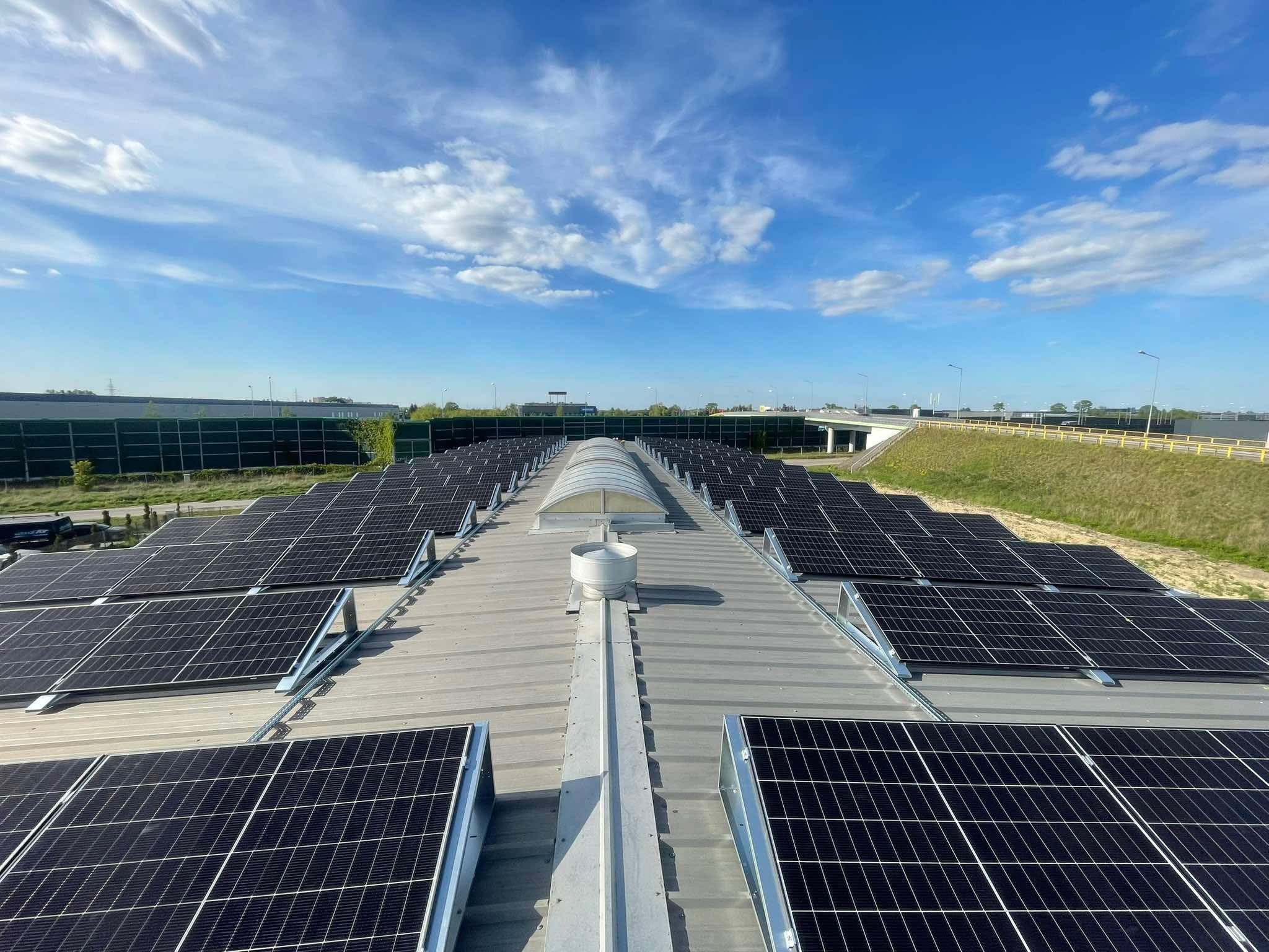 Instalacja solarna na dachu przemysłowym z świetlikiem i wentylacją, w tle błękitne niebo z chmurami i droga szybkiego ruchu.
