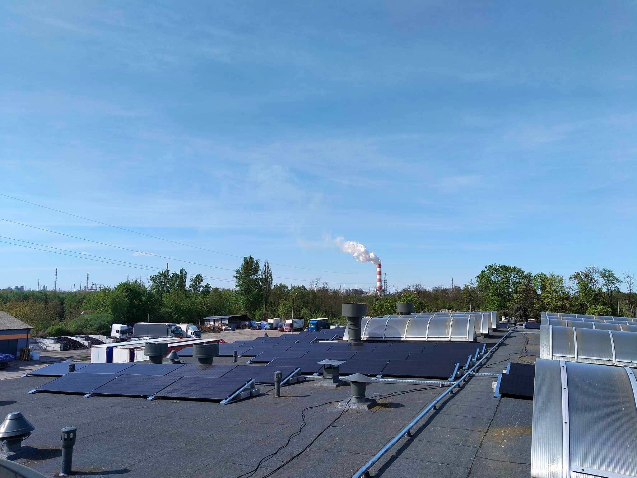 Instalacja paneli na dachu przemysłowym z kominem w tle. Widok z góry, błękitne niebo. Czarny dach z panelami i świetlikami.
