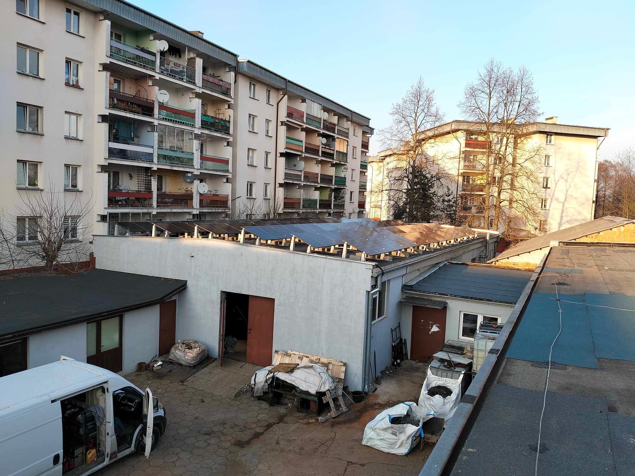 Montaż paneli na dachu budynku gospodarczego, widok z góry na częściowo zamontowaną instalację. W tle bloki mieszkalne i błękitne niebo.