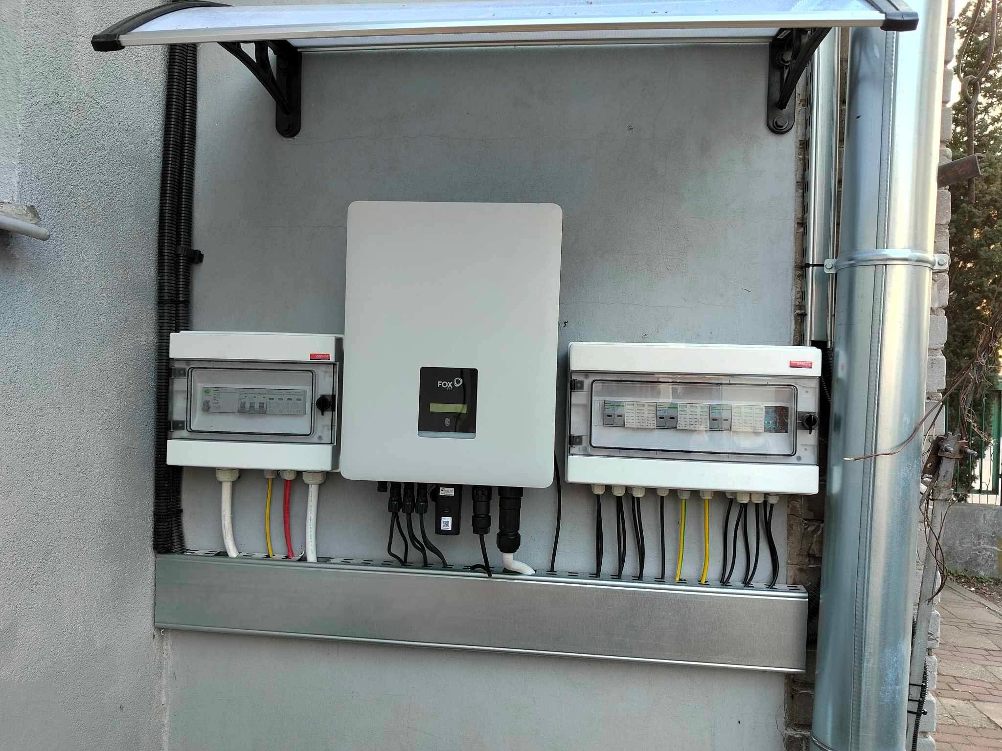 Zamontowany inwerter fotowoltaiczny marki FOX z rozdzielnicami na szarej ścianie budynku, z widocznymi przewodami i rurą spustową. Instalacja pod zadaszeniem.