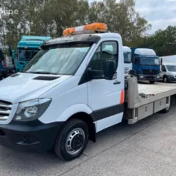 Biała laweta Mercedes Sprinter z pomarańczowym światłem ostrzegawczym na dachu, zaparkowana wśród innych pojazdów transportowych. Widok z przodu, lekki skos.