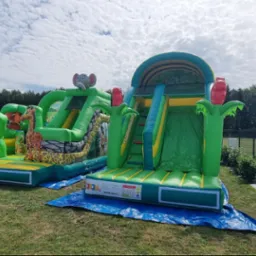 Dwa dmuchańce w stylu safari na trawie: jeden z motywem lwa i żyrafy, drugi ze słoniem. Tło to drzewa i błękitne niebo z chmurami.