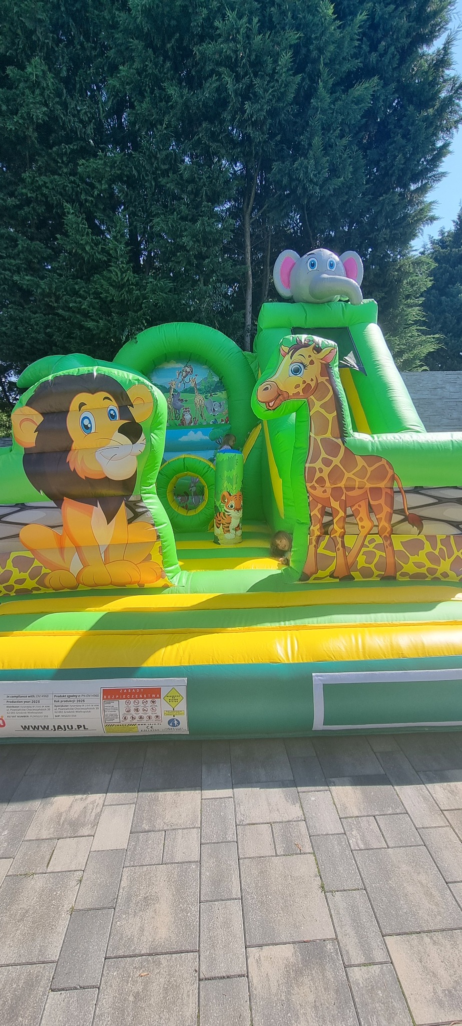 Kolorowy dmuchany zamek w motywie safari z lwem, żyrafą i słoniem. Widoczne drzewa w tle. Na zamku bawią się dzieci. Atrakcja dla dzieci na imprezę plenerową.