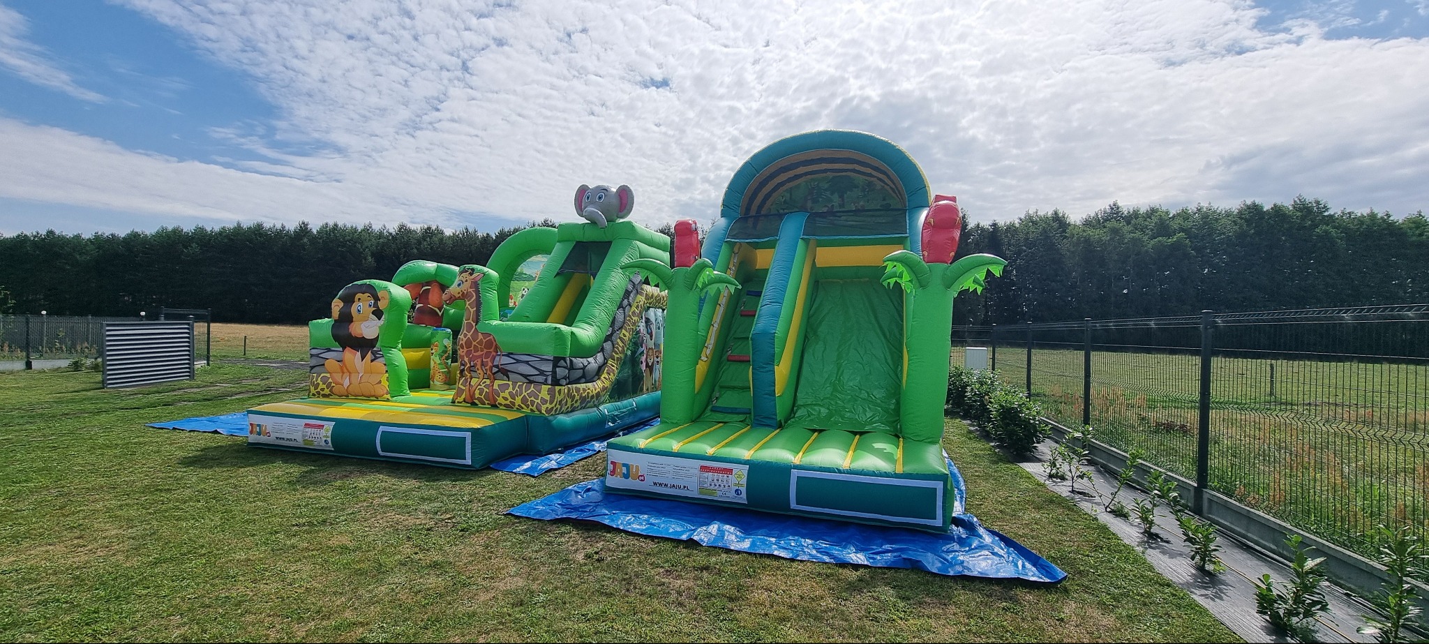 Dwa dmuchańce w stylu safari na trawie: jeden z motywem lwa i żyrafy, drugi ze słoniem. Tło to drzewa i błękitne niebo z chmurami.