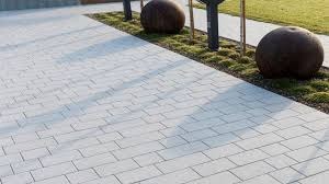 Jasnoszara kostka brukowa ułożona we wzór jodełki, z widocznymi kulami dekoracyjnymi i zielenią w tle. Delikatne cienie podkreślają strukturę kostki.