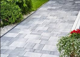 Szara kostka brukowa ułożona w geometryczny wzór, tworząca ścieżkę w ogrodzie. Po bokach roślinność, w tym kwitnące na czerwono kwiaty w donicy.