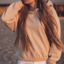 ZMATRYCY Paweł Niedźwiedź - Portret kobiety w beżowej bluzie z kapturem i białych spodniach na tle plaży o zachodzie słońca. Długie, proste włosy opadają jej na ramiona.