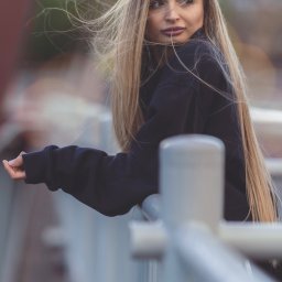 ZMATRYCY Paweł Niedźwiedź - Portret kobiety z długimi blond włosami, opierającej się o metalową barierkę na tle rozmytego krajobrazu miejskiego. Ujęcie z profilu, w swobodnej pozie.