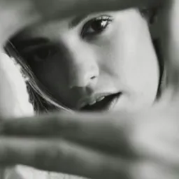 Czarno-biały portret kobiety z bliska, z delikatnym uśmiechem, widzianej przez nieostre przeszkody, tworzące unikalną ramę dla twarzy. Oświetlenie podkreśla rysy.