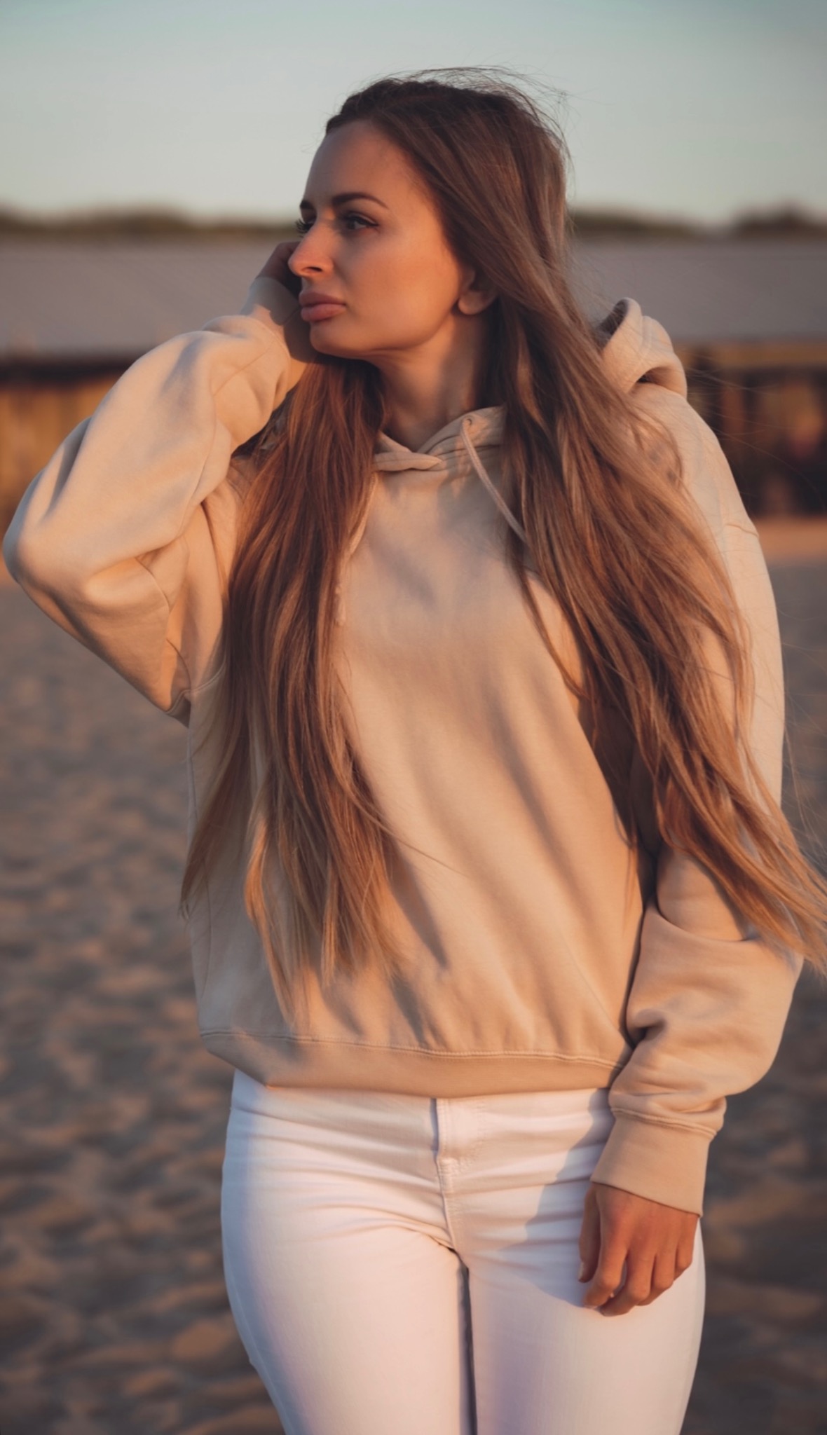 Portret kobiety w beżowej bluzie z kapturem i białych spodniach na tle plaży o zachodzie słońca. Długie, proste włosy opadają jej na ramiona.