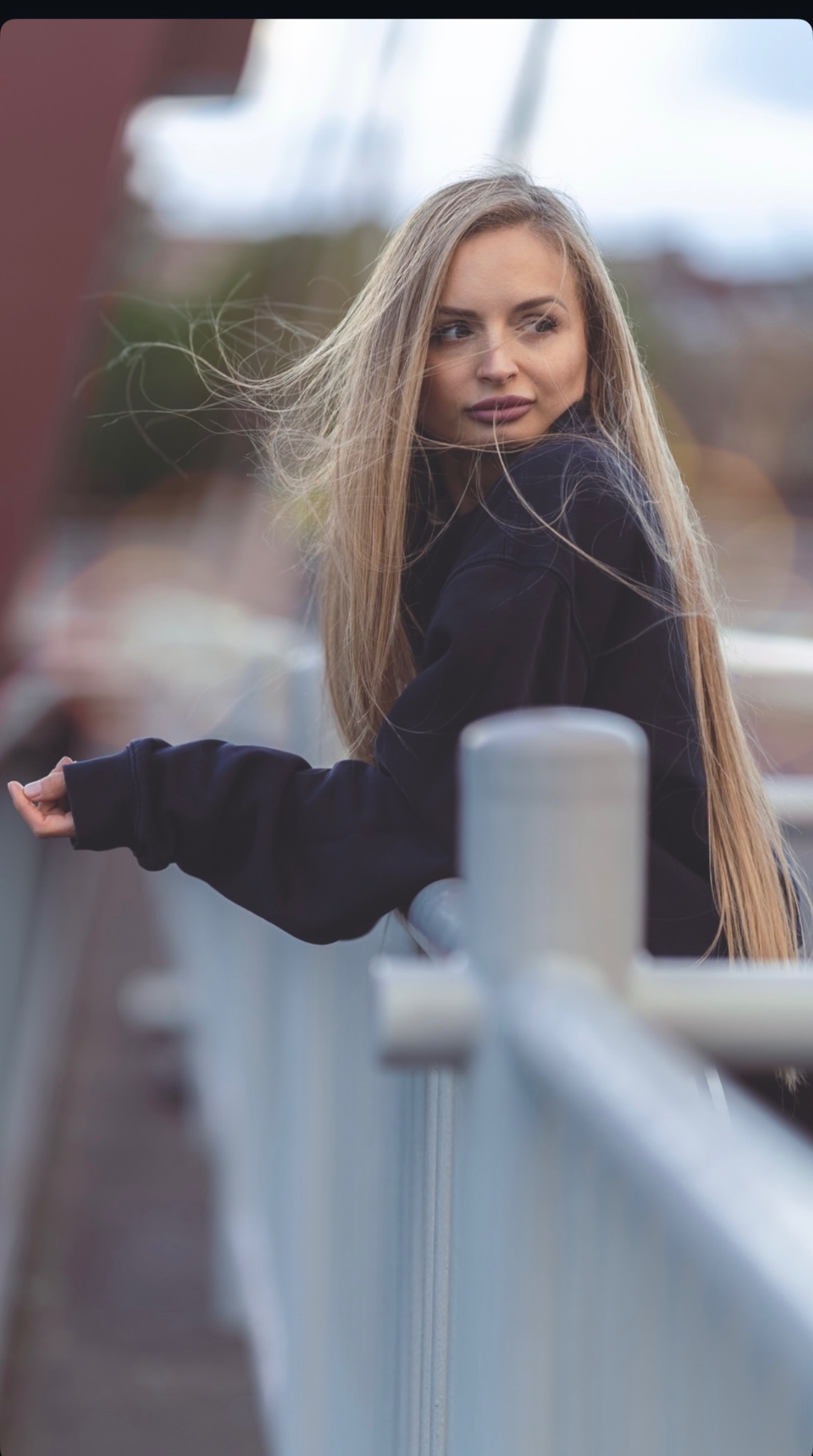 Portret kobiety z długimi blond włosami, opierającej się o metalową barierkę na tle rozmytego krajobrazu miejskiego. Ujęcie z profilu, w swobodnej pozie.