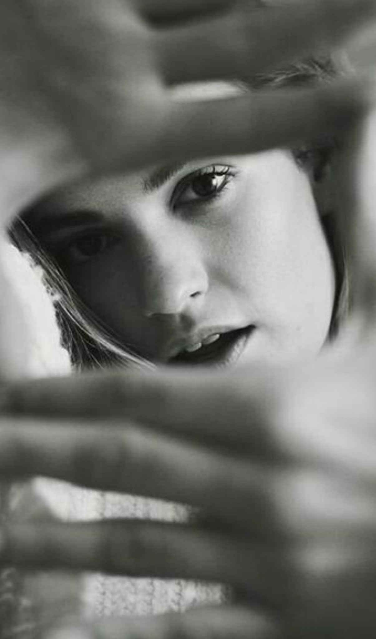 Czarno-biały portret kobiety z bliska, z delikatnym uśmiechem, widzianej przez nieostre przeszkody, tworzące unikalną ramę dla twarzy. Oświetlenie podkreśla rysy.