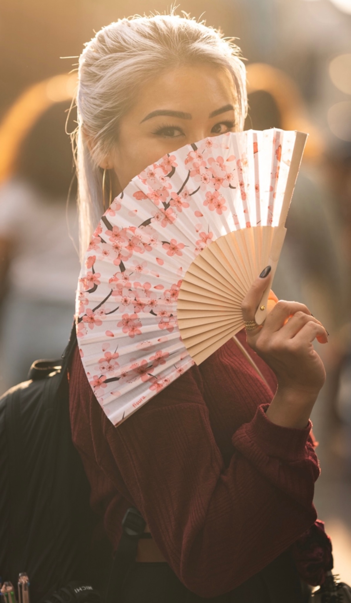 Portret kobiety z wachlarzem w kwiaty wiśni, ukrywającej część twarzy. Widoczne oczy, blond włosy i dłoń z ciemnym manicure w ciepłym oświetleniu.