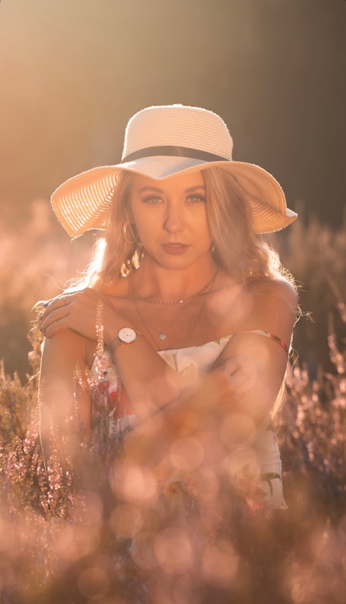 Portret kobiety w kapeluszu na tle kwitnącego wrzosowiska, złociste oświetlenie, delikatne rozmycie pierwszego planu, naturalny makijaż, subtelna biżuteria.