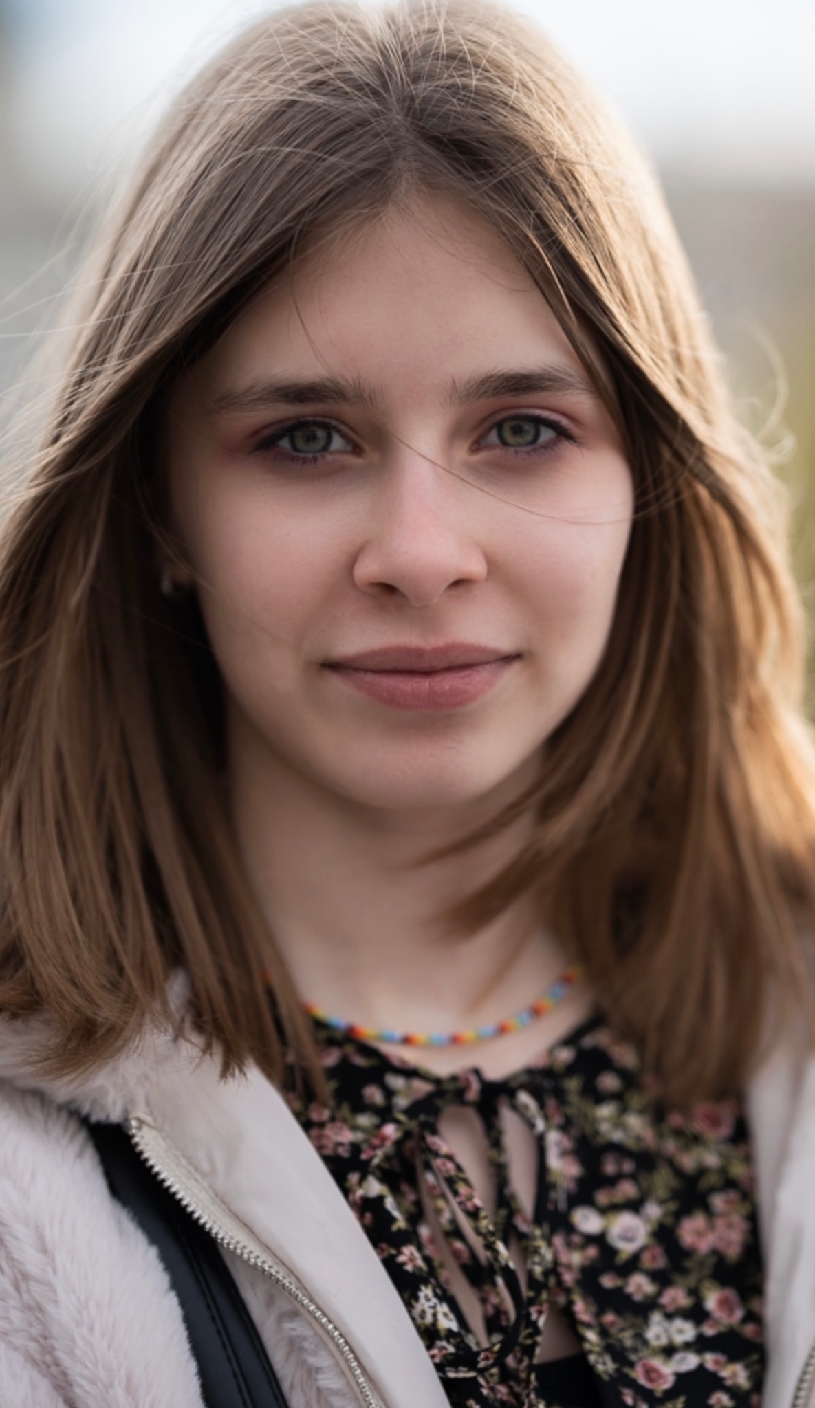 Portret młodej kobiety z długimi, brązowymi włosami i zielonymi oczami, ubranej w kwiecistą sukienkę i jasną kurtkę, z delikatnym uśmiechem.