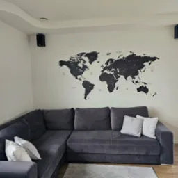 Nowoczesny salon z szarą sofą narożną, czarną mapą świata na ścianie i głośnikami. Minimalistyczny design i jasne wnętrze z drewnianą podłogą.