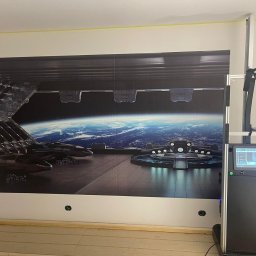 BaseElektric Artur Idasiak - Wielkoformatowy druk na ścianie z motywem futurystycznego hangaru kosmicznego. Widoczny automat drukujący i fragment drewnianej podłogi. Żółta taśma malarska przy suficie.