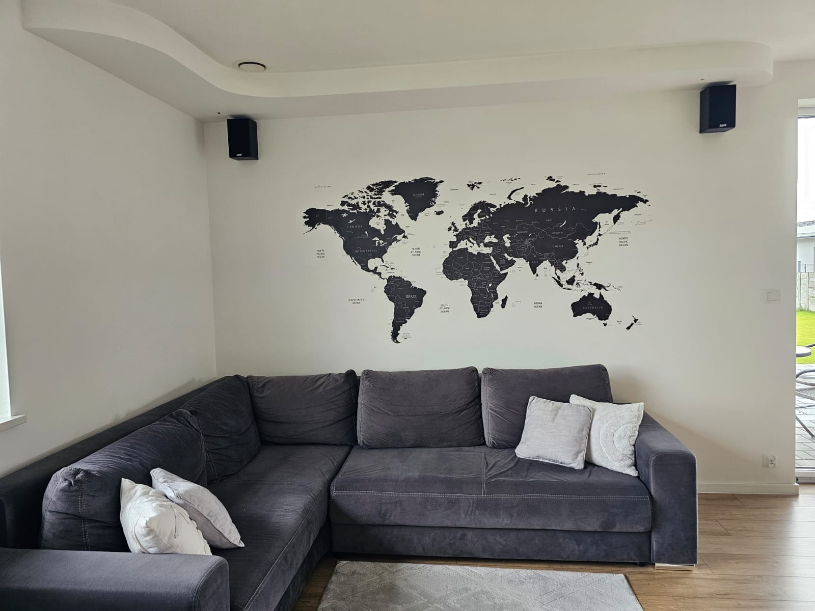 Nowoczesny salon z szarą sofą narożną, czarną mapą świata na ścianie i głośnikami. Minimalistyczny design i jasne wnętrze z drewnianą podłogą.