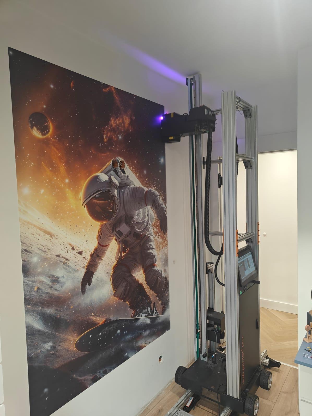 Drukarka ścienna w trakcie nanoszenia grafiki z motywem kosmicznym na ścianę w pomieszczeniu. Widoczny astronauta na desce snowboardowej na tle planet i gwiazd.