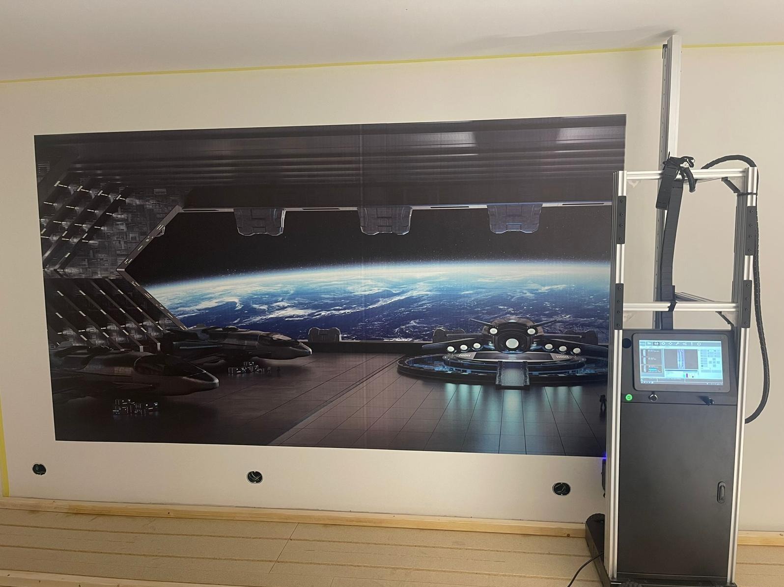 Wielkoformatowy druk na ścianie z motywem futurystycznego hangaru kosmicznego. Widoczny automat drukujący i fragment drewnianej podłogi. Żółta taśma malarska przy suficie.