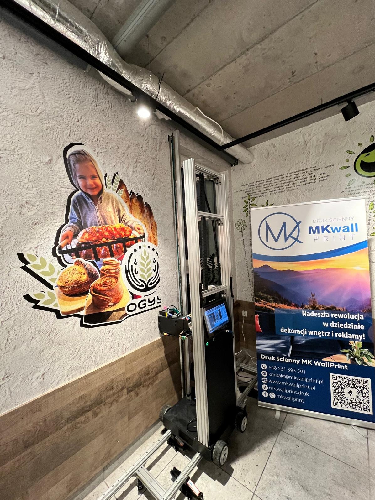 Drukarka ścienna MK WallPrint w trakcie pracy, nanosi kolorową grafikę z motywem piekarniczym na ścianę o chropowatej fakturze w lokalu gastronomicznym.