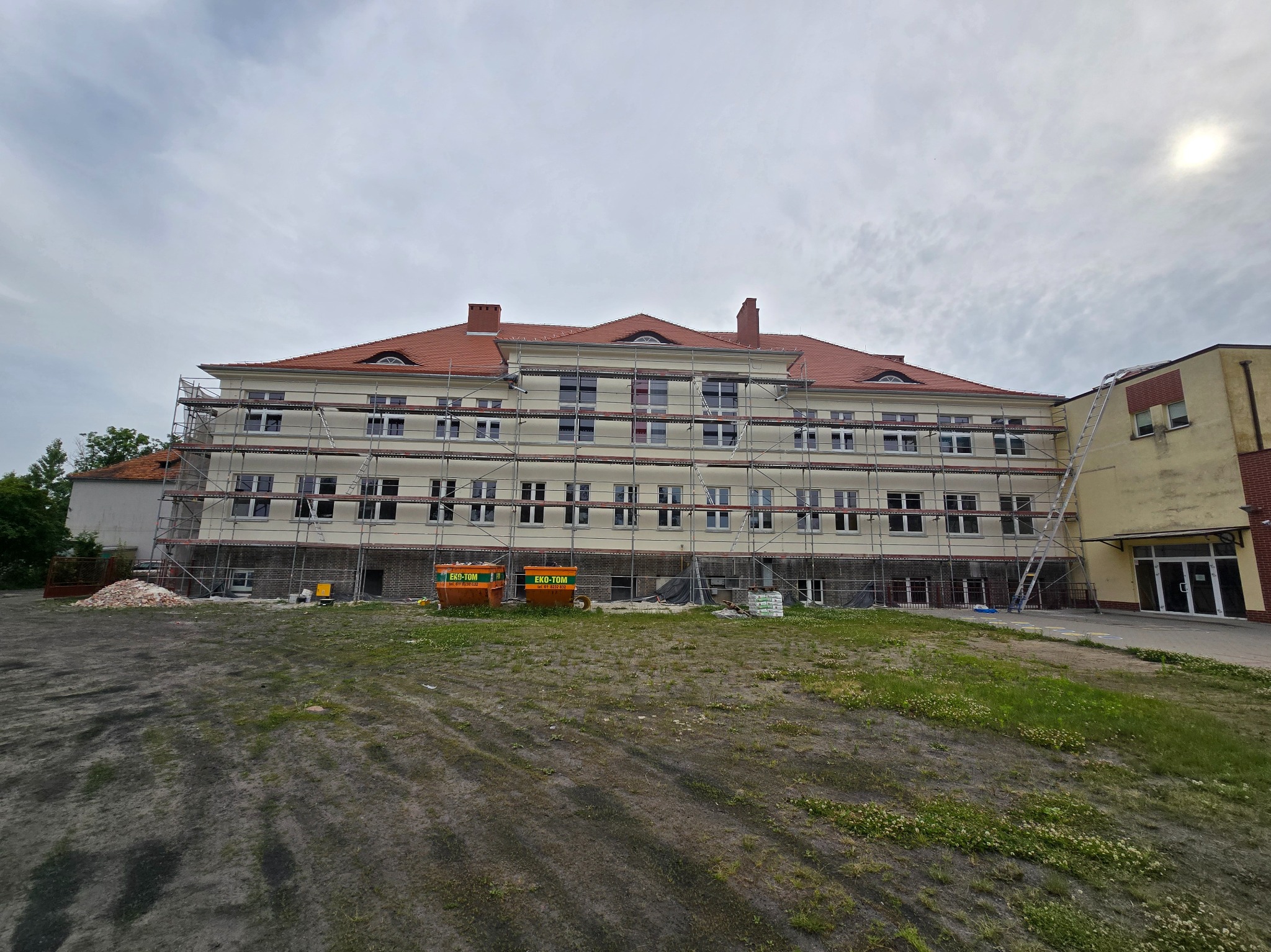 Budynek w trakcie malowania elewacji, otoczony rusztowaniami, z widocznymi kontenerami na odpady budowlane i stertą cegieł. Prace w toku, pochmurne niebo.