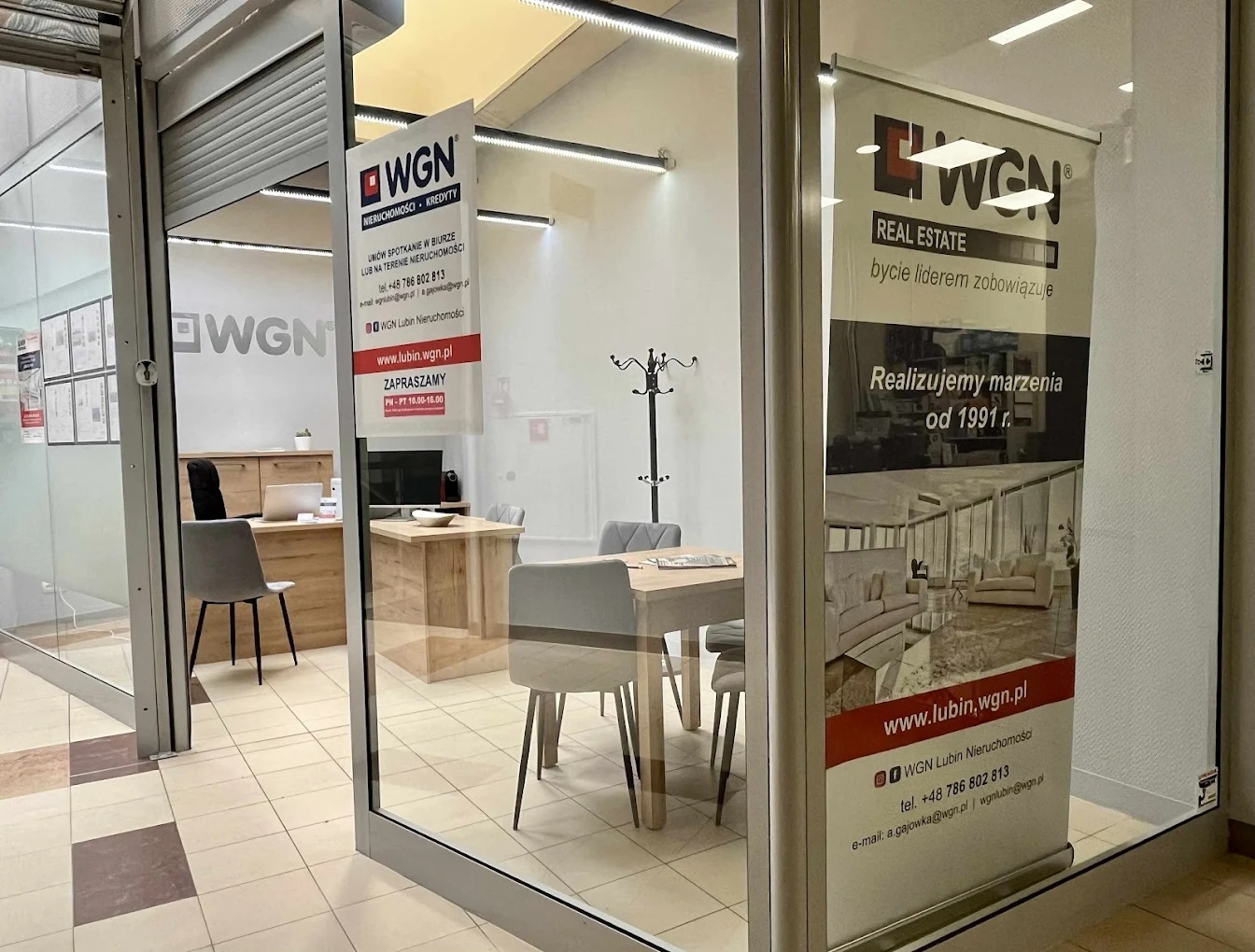 Wnętrze biura WGN Lubin Nieruchomości za przeszklonym wejściem, z plakatem reklamowym i informacjami kontaktowymi na szybie.