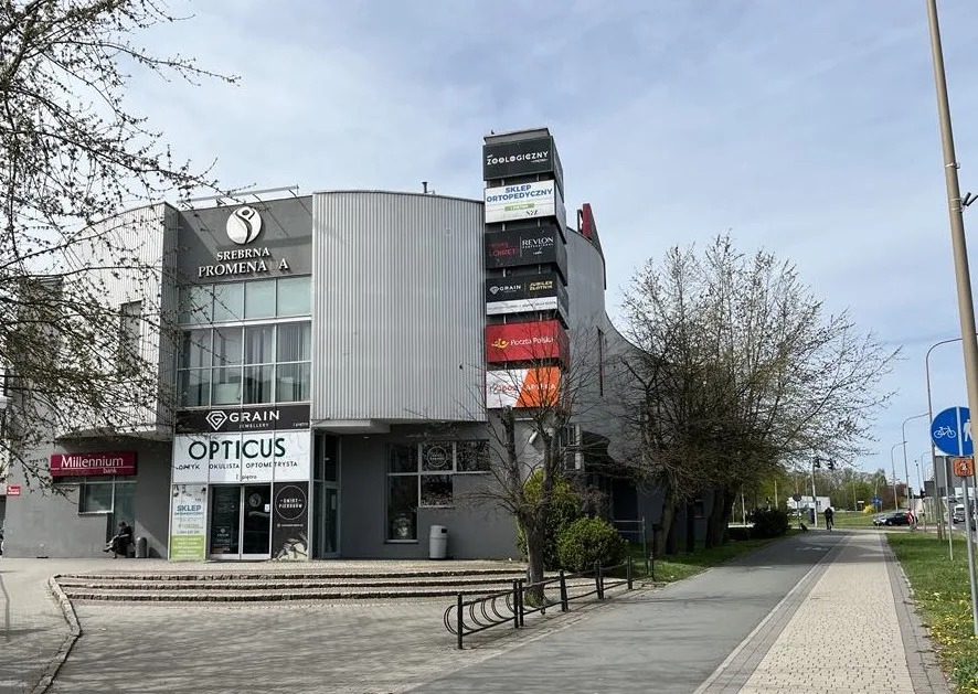 Fasada budynku Srebrna Promenada A z szyldami sklepów: zoologiczny, optyk, ortopedyczny, jubiler, Poczta Polska. Widok z chodnika z drzewami i ścieżką rowerową.