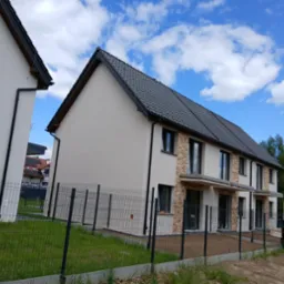 Nowoczesny, szeregowy dom w jasnej kolorystyce z elementami kamiennej elewacji, ogrodzony metalową siatką na tle błękitnego nieba z chmurami.