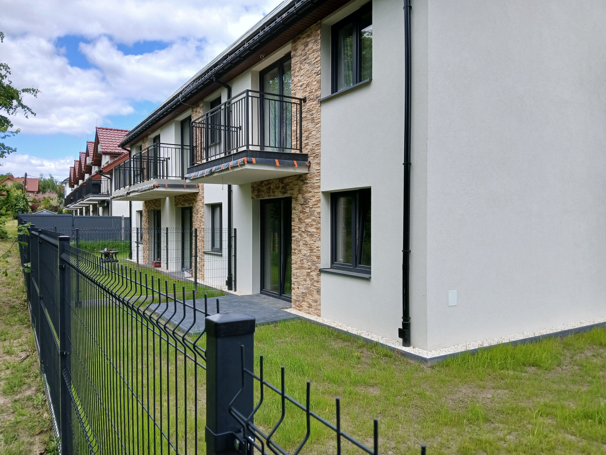 Rząd nowych domów z balkonami i elewacją w odcieniach bieli i kamienia, ogrodzony czarnym metalowym płotem na tle zielonego trawnika i błękitnego nieba.