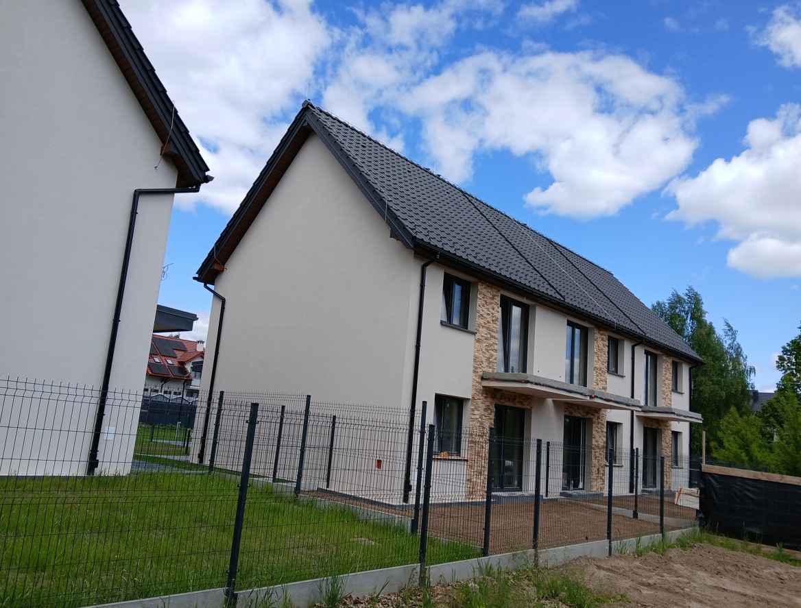 Nowoczesny, szeregowy dom w jasnej kolorystyce z elementami kamiennej elewacji, ogrodzony metalową siatką na tle błękitnego nieba z chmurami.