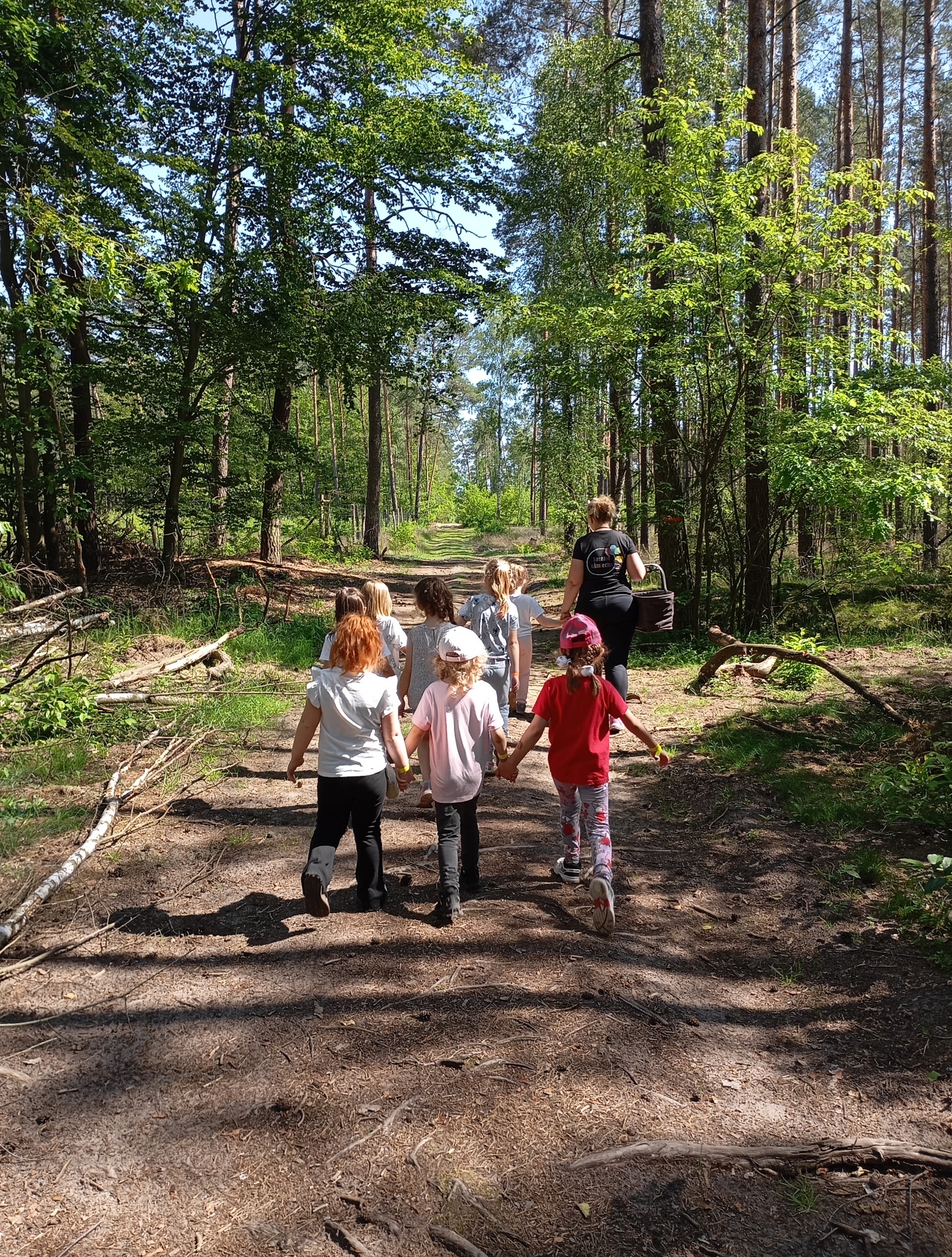 Grupa dzieci z opiekunem na leśnym spacerze, trzymają się za ręce. Widok z tyłu, słoneczny dzień.