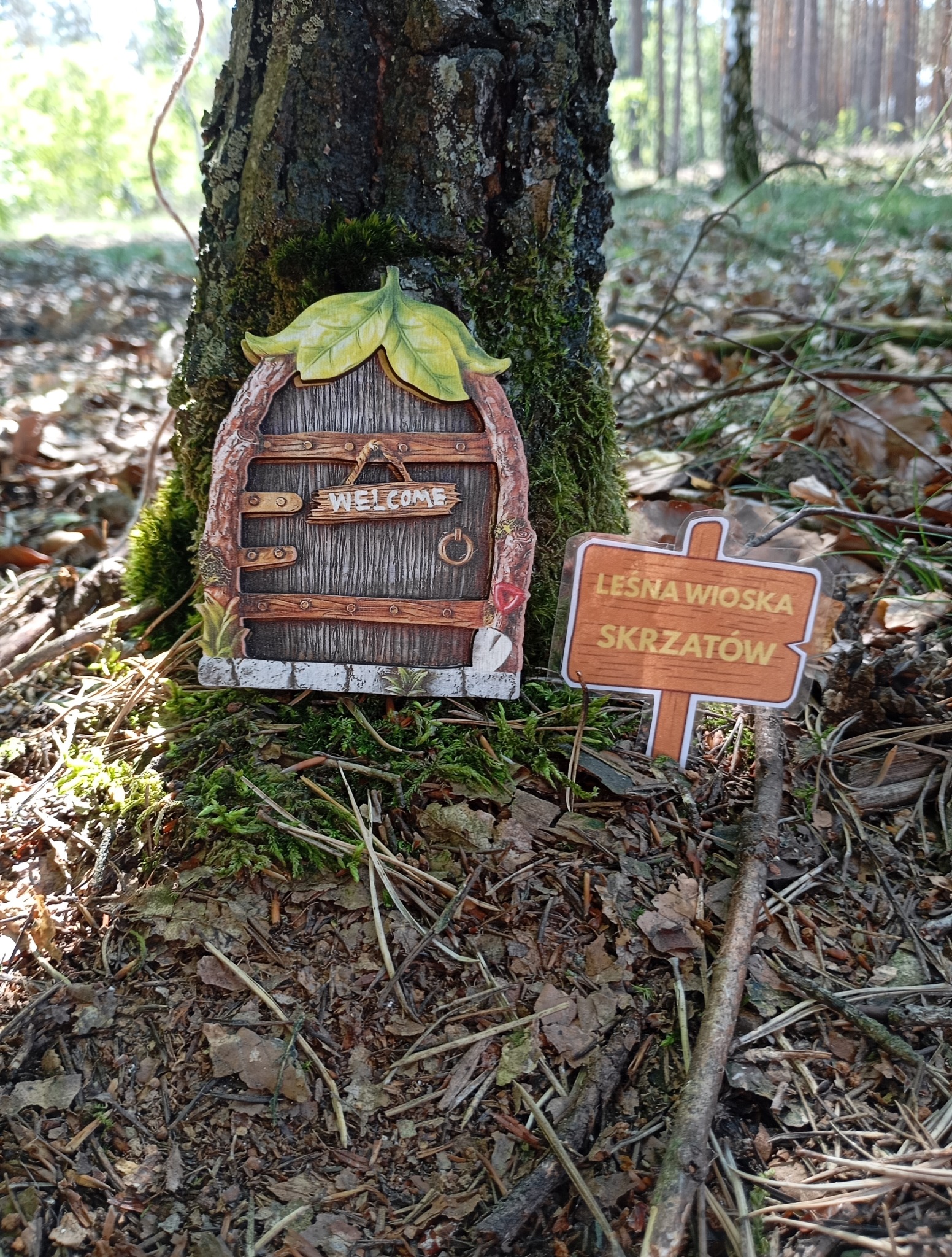 Dekoracyjna, miniaturowa wioska skrzatów z drewnianymi drzwiczkami i tabliczką informacyjną, umieszczona u podnóża drzewa w leśnym otoczeniu.