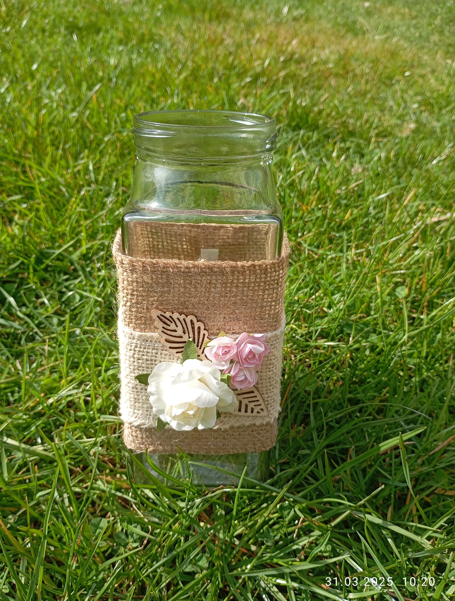 Szklany wazon zdobiony jutą i kwiatami, leżący na trawie. Dekoracja w stylu rustykalnym, idealna na przyjęcia okolicznościowe na świeżym powietrzu.