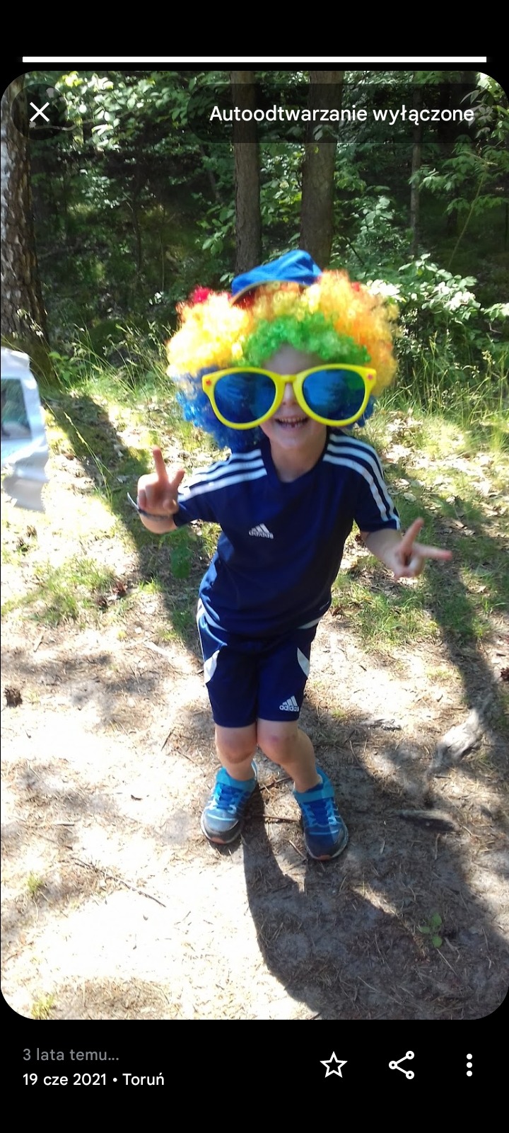 Uśmiechnięty chłopiec w kolorowej peruce i dużych okularach, w lesie, pokazuje znak zwycięstwa, w tle drzewa. Kostium imprezowy.