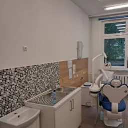 Umywalka z mozaiką w gabinecie dentystycznym, z niebieskim fotelem dentystycznym w tle. Widok na okno i zieleń na zewnątrz. Dominuje biel i jasne kolory.