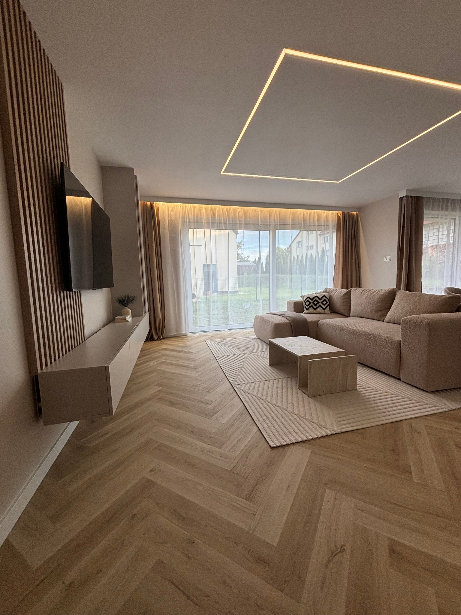 Jasny salon z parkietem w jodełkę, beżową sofą i podświetlanym sufitem. Minimalistyczny design, duże okno, naturalne światło. Dekoracyjne lamele na ścianie.