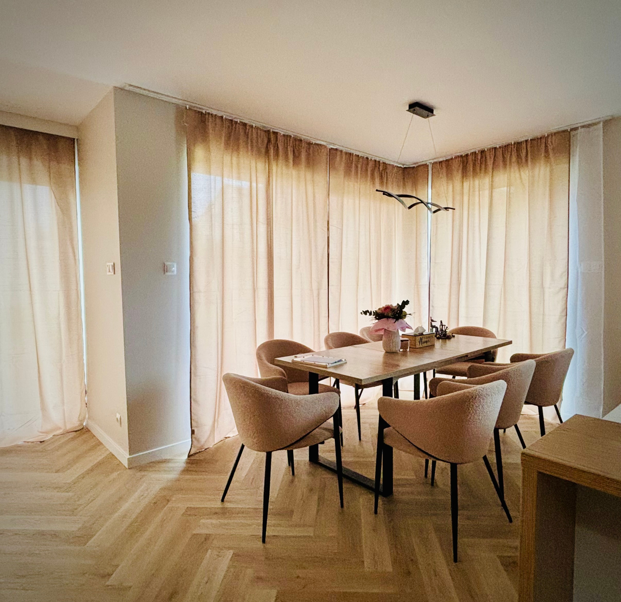Jasne wnętrze jadalni po remoncie: parkiet w jodełkę, beżowe zasłony, drewniany stół z kwiatami i nowoczesna lampa. Widok na wykończone pomieszczenie.